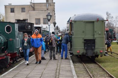 Grupa ludzi opuszcza stary parowóz i zielony wagon kolejowy stojący na wąskotorowej linii kolejowej. Osoby w różnorodnym zimowym ubiorze rozmawiają i pozują do zdjęć. Otoczenie jest klarowne, a tory kolejowe prowadzą w głąb kadru, gdzie widać kolejne budynki.