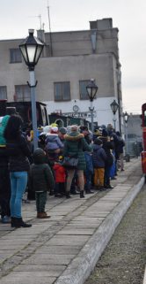 Grupa osób stoi na peronie w oczekiwaniu na czerwony wagon kolejki wąskotorowej, który znajduje się w odległości od tłumu. Na pierwszym planie widać tory i latarnię uliczną. W tle widać budynek i inne fragmenty infrastruktury kolejowej.