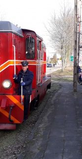 Czerwono-biały wąskotorowy pociąg stoi na torach obok peronu; podejrzewam, że jest to lokomotywa spalinowa. Po prawej stronie widoczny jest zielony wagon pasażerski. Na peronie dostrzec można kilka osób, w tym co najmniej jedno dziecko.