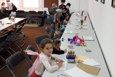 Dzieci w różnych kostiumach siedzą przy długim stole z kolorowymi kartkami i przyborami do rysowania. Jedno z dzieci ma różowe skrzydełka, a inne noszą kolorowe czapeczki. W tle dorosłe osoby towarzyszą dzieciom, jedna z nich również zajmuje się twórczą pracą przy stole.