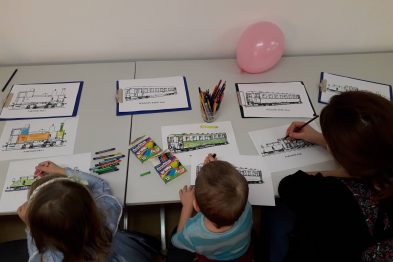 Dzieci siedzą przy stole i kolorują obrazki z motywami kolejowymi. Ubrani są w codzienne ubrania, skupieni na zadaniu z kredkami w rękach. W tle widać białą ścianę i różowy balon.