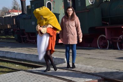 Dwoje ludzi idzie obok siebie po peronie kolejowym. Osoba z lewej jest przebrana za postać z dużą, żółtą głową i czerwonym strojem, trzyma maskotkę i jest zwrócona twarzą w kierunku kamery. Osoba z prawej, w okularach i różowym płaszczu, jest zwrócona w stronę towarzysza i uśmiecha się, w tle widać zielone lokomotywy parowe.