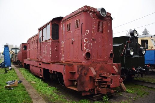 Czerwona lokomotywa spalinowa stoi na torach; jest to model typowy dla kolei wąskotorowych. Wygląda na starszy typ i wymaga remontu. W otoczeniu lokomotywy widoczne są inne pojazdy kolejowe i fragmenty infrastruktury kolejowej.