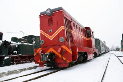 Czerwony wagon kolejowy stoi na zaśnieżonych torach obok innych pojazdów szynowych. Pomimo śniegu, sprzęt wygląda na sprawny i gotowy do jazdy. Perony i otoczenie są pokryte białym puchem, co nadaje scenerii zimowy nastrój.