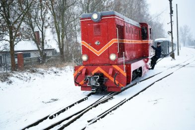 Czerwony pociąg retro stoi na pokrytych śniegiem torach; otoczenie wydaje się być zimowe z widocznymi opadami śniegu. Lokomotywa zdaje się być w dobrym stanie, jest zadbana i połyskująca. W tle widać drzewa i niewielkie budynki przykryte białym puchem.