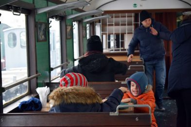 W drewnianym wnętrzu retro wagonu kolejowego siedzą pasażerowie; dzieci i dorośli ubrani w zimowe odzieże. Jeden z dzieci uśmiecha się, a dorosły w tle stoi i trzyma się poręczy. Przez okno wagonu widać wagony stojące na zewnątrz i pokryte śniegiem tory.