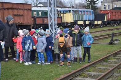 Grupa dzieci stoi na peronie kolejowym, otoczona przez historyczne wagony kolejowe i torowisko. Dzieci są ubrane w ciepłe kurtki i czapki, a niektóre z nich mają plecaki. W tle widać zabytkowe wagony towarowe i osobowe w różnych kolorach.
