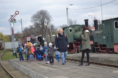 Grupa dzieci prowadzona przez dorosłych idzie wzdłuż torów kolejowych, gdzie stoją stare zielone lokomotywy parowe. Na tle pochmurnego nieba widać zabytkowe wagony kolejowe i semafor. Dzieci są ubrane w kolorowe kurtki i czapki, a niektóre z nich trzymają w rękach zabawki lub małe plecaki.
