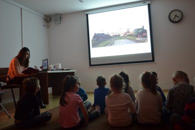 Grupa dzieci siedzi na podłodze skupiona na obrazie prezentowanym przez rzutnik, przedstawiającym tory kolejowe. Osoba dorosła siedzi obok nich, opowiadając i wskazując na ekran. Cała scena ma miejsce w pomieszczeniu z wyjątkiem dzieci, które mają na sobie kolorowe ubrania.