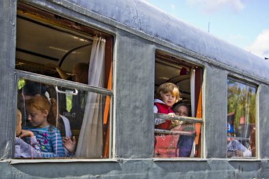 W oknach starego wagonu kolejowego widoczni są pasażerowie, wśród których są dzieci i dorośli. Wagon ma metalowe ramy okien i białe zasłony. Osoby siedzące wewnątrz patrzą na zewnątrz z zainteresowaniem.