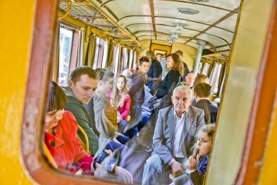Pasazerowie siedza i stoja w zabytkowym wagonie kolejowym; dzieci i dorośli wyglądają przez okna lub zajmują się sobą. Wagon ma drewniane wykończenia i utrzymany jest w stylu retro. Światło wpadające przez okna tworzy jasną i przyjazną atmosferę.