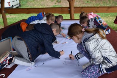 Grupa dzieci siedzi przy długim stole, na którym rozłożony jest biały arkusz do rysowania. Dzieci są skupione na rysowaniu lub pisaniu, używając kolorowych kredek i flamastrów. W tle widać namiot i zieloną roślinność, co wskazuje na plenerowe warunki.