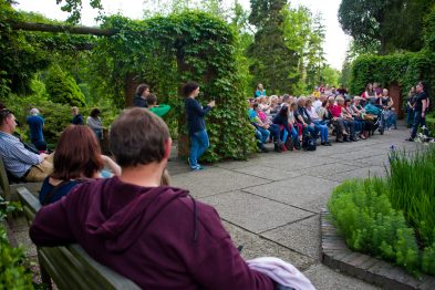 Grupa osób siedzi na ławkach i wsłuchuje się w prezentację, którą prowadzi osoba stojąca przed nimi. Publiczność jest skoncentrowana, otaczają ich zielone rośliny i długie żywopłoty w zacisznym ogrodzie. Wydarzenie odbywa się w jasny dzień, a uczestnicy noszą casualowe ubrania.