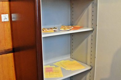 Metalowa szafa archiwizacyjna jest otwarta, ukazując cztery półki na dokumenty i przedmioty. Na dwóch górnych półkach znajdują się segregatory i papiery, a dwie dolne są puste. Obok szafy widać część drewnianej powierzchni mebla, być może biurka lub szafki.