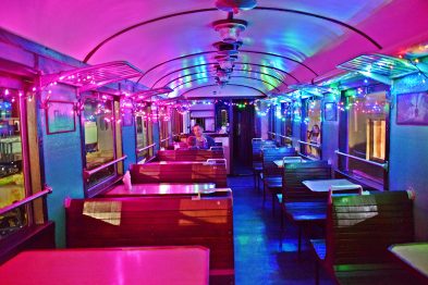 Wagon restauracyjny oświetlony kolorowymi światłami tworzy wyjątkową atmosferę dla odwiedzających. Stoliki przygotowane są do przyjęcia gości, a na ścianach widać dekoracyjne elementy i obrazy. Podłoga jest czysta i lśniąca, co dodaje wnętrzu elegancji.