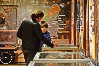 Dwóch mężczyzn w muzeum ogląda eksponaty kolejowe umieszczone w szklanych witrynach; jeden z nich wskazuje na jakiś przedmiot. W tle widoczne są duże zdjęcia i mapy dotyczące historii kolei. Całe pomieszczenie jest dobrze oświetlone, co pozwala zwiedzającym na dokładne przyjrzenie się zgromadzonym artefaktom.