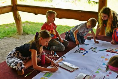 Grupa dzieci w różnym wieku siedzi przy długim stole, koncentrując się na rysowaniu i kolorowaniu na arkuszach papieru. Siedzą w drewnianej altanie, która zapewnia cień i schronienie. Różnokolorowe kredki, flamastry oraz arkusze papieru zajmują większość przestrzeni na stole.