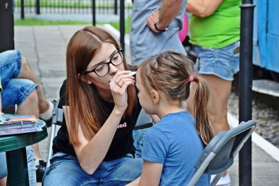 Kobieta w okularach maluje dziecku twarz podczas wydarzenia plenerowego. Siedzą one przy składanym stoliku, w tle widoczny jest wagon kolejowy. Uczestnicy eventu w tle zajęci są różnymi aktywnościami.