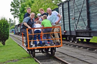 Grupa osób, w tym dorośli i dzieci, stoi na otwartej platformie kolejowej, uśmiechając się i pozując do zdjęcia. Obok platformy znajduje się zielony wagon towarowy. Tło stanowi dobrze utrzymany teren kolejowy z zielenią i szynami.