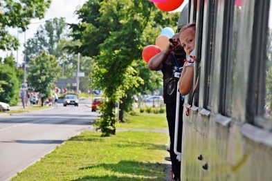 Dziecko wystawia głowę przez okno pociągu, trzymając w ręku kolorowe balony. Drzwi wagonu są otwarte, a za pojazdem widać drzewa i część chodnika. Słoneczna pogoda i zielone otoczenie dodają uroku scenie.