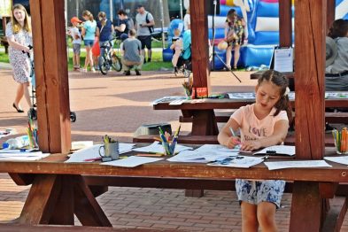 Dziewczynka koncentruje się na rysowaniu przy drewnianym stole z rozłożonymi kartkami i kredkami. W tle widać grupy ludzi, w tym dzieci, bawiących się na zewnątrz przy dmuchanych zamkach i innych atrakcjach. Atmosfera wydarzenia wydaje się radosna i rodzinna, z aktywnościami dla najmłodszych uczestników.