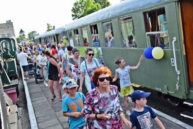 Na fotografii widoczni są ludzie, w tym dzieci i dorośli, na peronie obok zielonego wagonu kolejowego. Niektórzy uczestnicy trzymają kolorowe balony i wydają się rozmawiać oraz czekać na odjazd pociągu. Pogoda jest ładna, a scena wydaje się być zarejestrowana podczas publicznego wydarzenia lub festynu związanego z koleją.