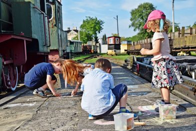 Grupa dzieci bawi się na peronie kolejowym malując kredą na betonowych płytach. Dziewczynka w różowym kapeluszu obserwuje, jak dwóch chłopców i jedna dziewczynka aktywnie rysują obok torowiska. W tle widoczne są zielone wagony kolejki wąskotorowej i jasne niebo.