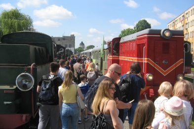 Ludzie gromadzą się na peronie wokół czerwonego lokomotywy wąskotorowej; wielu z nich to rodziny z dziećmi. Słońce świeci i jest to dzień o jasnym niebie. Tłum odbywa podróż w stylu retro, celebrując Dzień Dziecka w muzeum kolei.