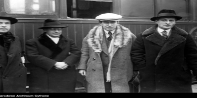 Czterech mężczyzn stoi obok siebie przed wagonem kolejowym; wszyscy są ubrani w ciepłe, eleganckie płaszcze i kapelusze typowe dla dawnych epok. Drugi od lewej mężczyzna wyróżnia się jasnym płaszczem i białym kapeluszem, podczas gdy pozostali noszą ciemniejsze ubrania. W tle widoczna jest ściana wagonu, co podkreśla kolejowy kontekst sceny.