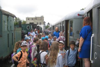 Grupa dzieci w wieku szkolnym, ubrana w letnie stroje i plecaki, zbiera się obok zielonych wagonów kolejowych. Dorosła przewodniczka w niebieskim stroju pomaga dzieciom przy wsiadaniu do pociągu. W tle widać maszynę parową, co nadaje scenie historyczny charakter.