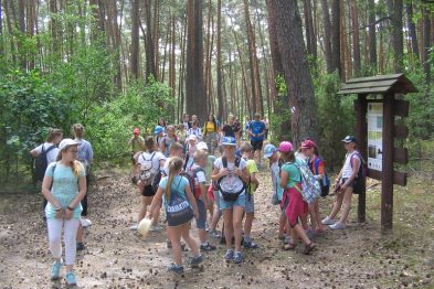 Grupa ludzi, w tym głównie dzieci i młodzież, stoi w lesie na ścieżce przy tablicy informacyjnej. Są ubrani w letnie stroje i większość z nich ma plecaki. W tle widać gęste drzewa i iglaste drzewostany.