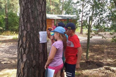 Dwie osoby, dorosły i dziecko, stoją przy drzewie, na którym zawieszona jest kartka z tekstem. Dziecko czyta informacje, trzymając kartkę w ręku, a dorosły wskazuje palcem na tekst, aby pomóc w zrozumieniu treści. W tle widoczne są drzewa i fragment drewnianego budynku, co sugeruje, że scena rozgrywa się na terenie zalesionym lub w parku.