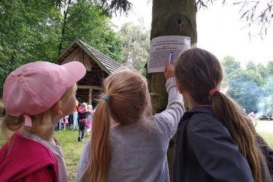 Trzy dzieci z zaciekawieniem czytają informacje zamieszczone na kartce przyczepionej do drzewa. Dwie dziewczynki w warkoczykach i chłopiec w kurtce są skupieni na tekście, którego nie widzimy. W tle zauważalna jest zielona przestrzeń z ludźmi i drewnianym budynkiem, co sugeruje plenerową aktywność edukacyjną.