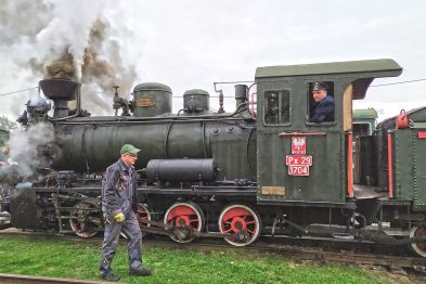 Parowóz oznaczony numerem Px29-1704 emituje dym, stoi na torach, a w kabinie widać maszynistę. Osoba ubrana w ciemną kurtkę i jasną czapkę przechodzi przed lokomotywą. W tle zauważalne są zielone drzewa i szara niebo.