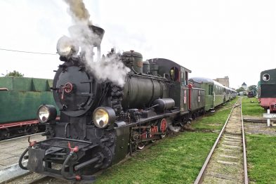 Parowóz Px29-1704 z wagonami stoi na torach wąskotorowych w otoczeniu zieleni i niskich budynków. Czarno-czerwona lokomotywa wydaje gęste strumienie białego dymu w górę. Sznur wagonów pasażerskich ciągnie się za parowozem, a obok biegnie równoległy tor kolejowy.