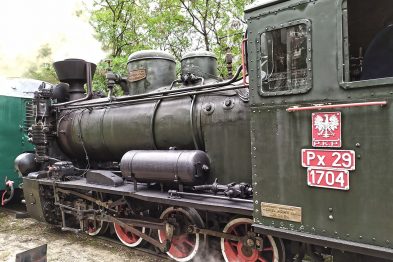 Czarny parowóz z czerwonymi obręczami na kołach i numerem Px29-1704 wypuszcza parę. Maszyna stoi na torach otoczona zielenią drzew. Tabliczka z białym tłem i czerwoną ramką przedstawia numer i typ lokomotywy.