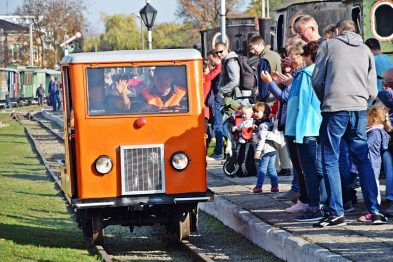 Niewielki pomarańczowy wagon kolejowy stoi na torze, otoczony grupą ludzi, w tym dziećmi. Jest słoneczny dzień, a tło obrazu stanowią zielone drzewa i inne elementy kolejowe. Odwiedzający rozmawiają ze sobą i widać, że niektórzy robią zdjęcia.