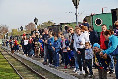 Grupa osób stoi na peronie obok torów, w tym dzieci i dorośli, niektórzy z nich robią zdjęcia. Za tłumem widać zieloną parowozownię i fragment starego parowozu z czarnym kominem. Atmosfera wydarzenia wydaje się przyjazna i pełna zainteresowania, a wszyscy skupiają uwagę na pociągu i torach.