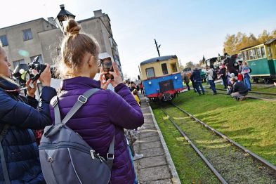 Ludzie z aparatami fotograficznymi robią zdjęcia stojącemu kolorowemu pociągowi wąskotorowemu; tłum zgromadził się obok torów. Na pierwszym planie widoczna jest kobieta trzymająca telefon komórkowy, a w tle niebo jest bezchmurne i jasne. Ogólne wrażenie jest radosne i ludzie wydają się być zainteresowani wydarzeniem.