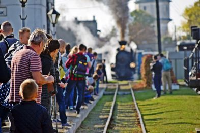Grupa osób stoi przy torach kolejowych, obserwując zbliżającą się parowóz z wypuszczającym dym kominem. Wśród nich są dorosłe osoby oraz dzieci, niektórzy są odwróceni tyłem do kamery. Słońce oświetla scenę, tworząc ciepłą atmosferę.