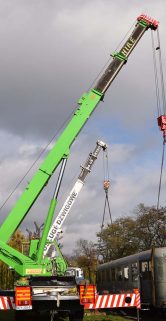 Podnośnik o zielonym ramieniu podnosi olbrzymi metalowy obiekt za pomocą czerwonego ucha haka, które jest przytwierdzone do środka konstrukcji. Urządzenie stoi na stabilnych, ustawionych poziomo podporach zabezpieczających działanie na terenie trawiastym i stabilizujących podnośnik podczas operacji. Metalowy obiekt, prawdopodobnym wagonem kolejowym, jest umieszczony w powietrzu nad trawiastą powierzchnią i wydaje się być transportowany lub przenoszony