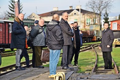 Grupa mężczyzn stoi na peronie kolejowym obok torów i starego wagonu; niektórzy z nich są zwróceni w stronę kamery. Jeden z mężczyzn ubrany w garnitur gestykuluje w trakcie rozmowy, podczas gdy reszta słucha. Tło stanowi budynek stacyjny oraz zielone drzewa.