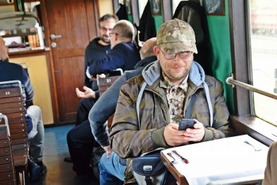 Grupa osób siedzi w drewnianym wnętrzu wagonu kolejowego, skupiając się na różnych aktywnościach. Jeden mężczyzna w centralnej części kadru, ubrany w kamuflażową kurtkę, patrzy na ekran telefonu komórkowego. Otaczają go inni uczestnicy, niektórzy rozmawiają, a jeden z nich trzyma dokumenty.