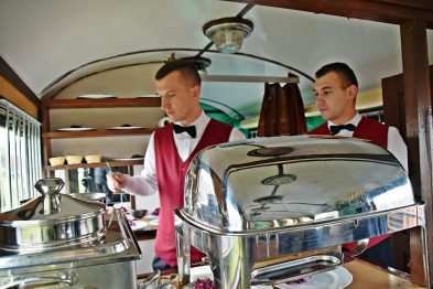 Dwaj mężczyźni w średnim wieku w eleganckich uniformach kelnerskich stoją przy bufecie na pokładzie wagonu restauracyjnego. Na metalowej ladzie widoczne są podgrzewane pojemniki na jedzenie. W tle prześwitują okna wagonu, przez które widać rozmazane kontury zewnętrznego środowiska.