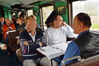 Grupa osób siedzi wewnątrz zabytkowego wagonu kolejowego, skupiona na rozmowach i słuchaniu prelekcji. Mężczyźni są ubrani w codzienne, nieformalne ubrania; jedna osoba trzyma dokumenty. W tle przez okna widać zieloną roślinność, sugerującą postój lub powolny ruch pociągu.