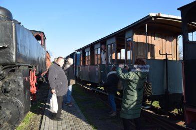 Lokomotywa parowa stoi na torach obok drewnianych wagonów. Ludzie są zwróceni w stronę pociągu; niektórzy rozmawiają i robią zdjęcia. Słońce świeci, a niebo jest bezchmurne, co wskazuje na pogodny dzień.