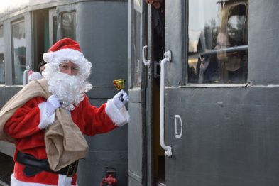 Osoba przebrana za Świętego Mikołaja stoi przy pociągu i dzwoni złotym dzwonkiem. Ma na sobie charakterystyczne czerwone ubranie z białym futerkiem oraz długą białą brodę. W tle widoczne są zarysy wagonu kolejowego oraz fragment torów.