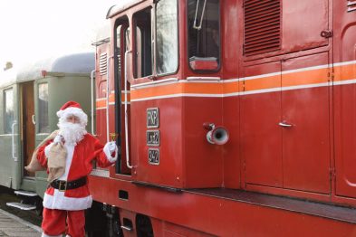 Osoba przebrana za Świętego Mikołaja stoi obok czerwonej lokomotywy wąskotorowej z wagonami w tle. Święty Mikołaj trzyma dzwonek w jednej ręce i macha do kamery drugą. Lokomotywa i wagony wyglądają na odrestaurowane i są zaparkowane na szynach.