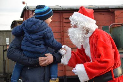 Dziecko w niebieskiej czapce i niebieskiej kurtce rozmawia z postacią ubraną w czerwony strój Mikołaja ze śnieżnobiałą brodą. Stoją przed czerwonym wagonem kolejki wąskotorowej. Atmosfera wydaje się być radosna i świąteczna.