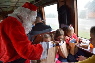 Osoba przebrana za Świętego Mikołaja rozdaje prezenty dzieciom siedzącym w drewnianym wnętrzu wagonu kolejowego. Dzieci z zainteresowaniem przeglądają zawartość papierowych torebek, a jeden z chłopców wyjmuje coś z torebki. Tło rozmyte wskazuje na ruch pociągu.